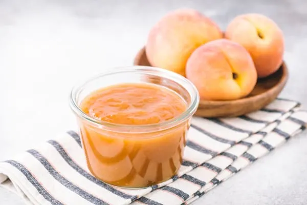 Vanilla honey peach butter in a glass jar sitting on a striped napkin with peaches in the background.