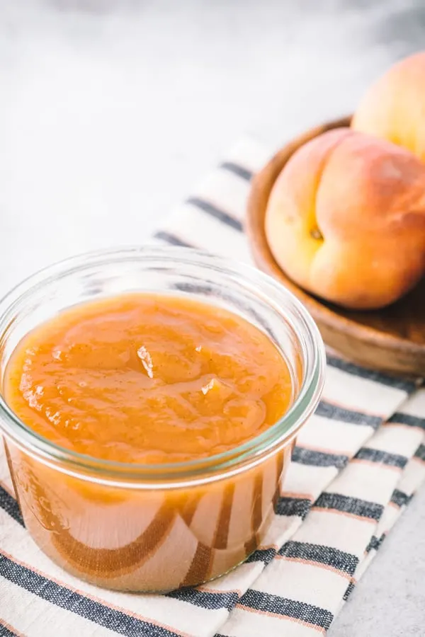 Small glass jar filled with vanilla honey peach butter with peaches in the background.