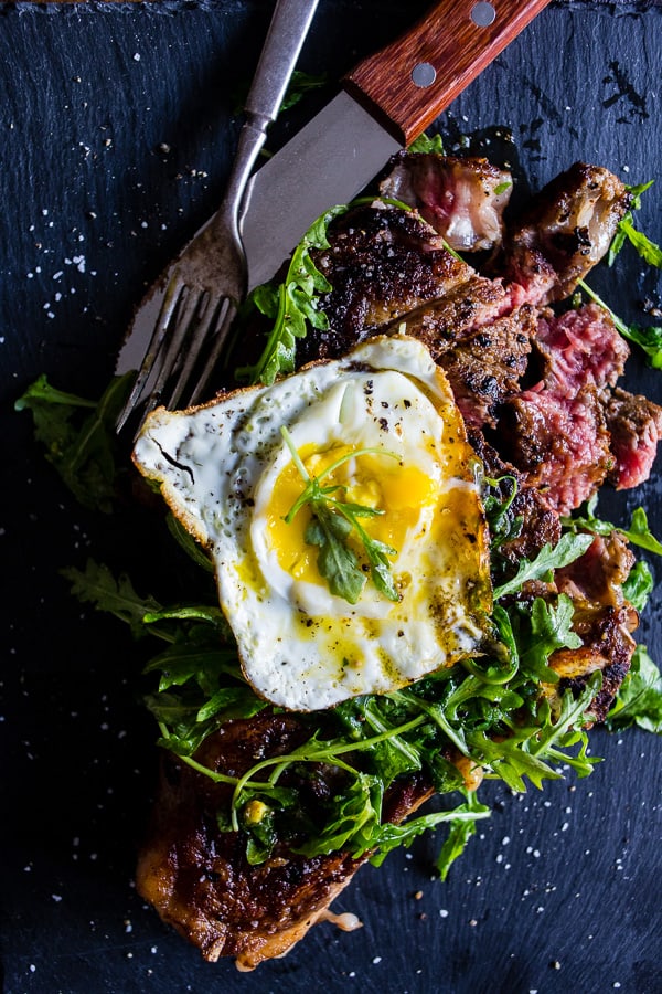 fried egg topped grilled ribeye steak with arugula salad sitting on a black plate