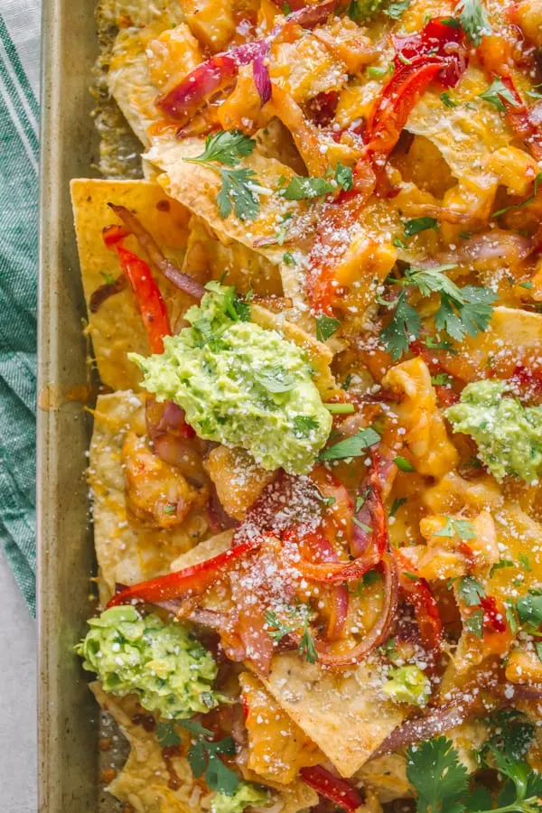 Close up of nachos on a sheet pan covered in chicken, cheese, onions, guacamole and cilantro. 