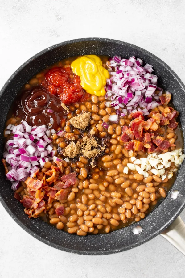 easy bacon baked beans in a skillet with all the ingredients on top before being mixed.