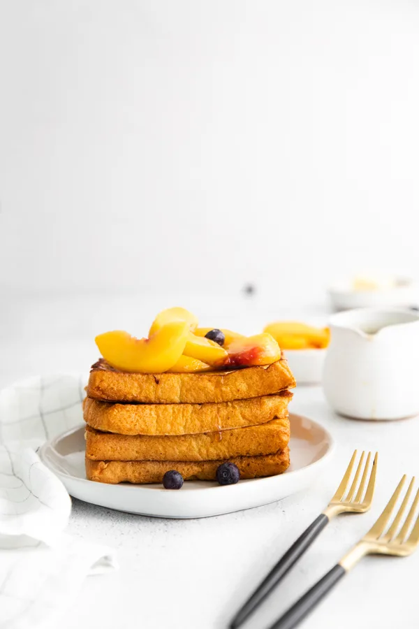 Peach Topped French Toast stacked on a plate topped with peaches and maple syrup.