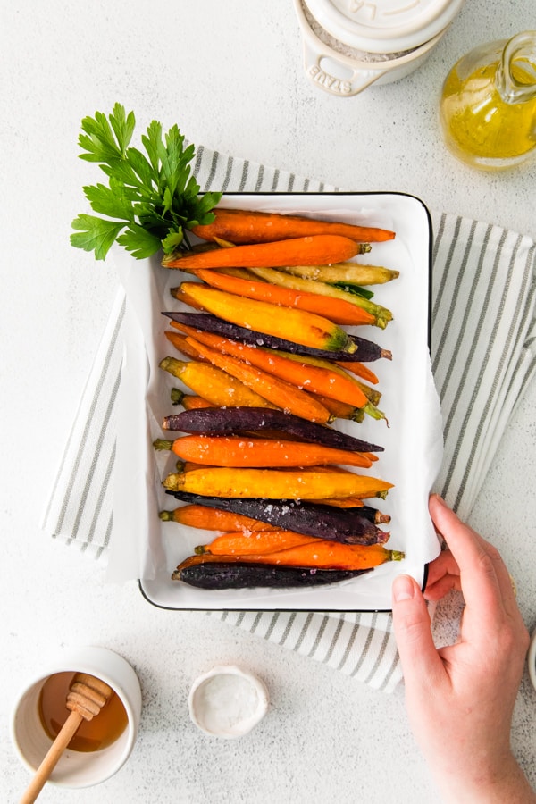 Honey Roasted Carrots cooking a serving dish with a hand touching the dish. 