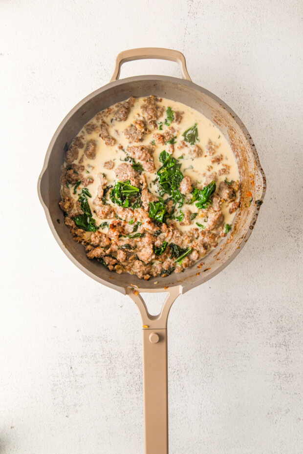Adding cream to a skillet with sausage and kale. 